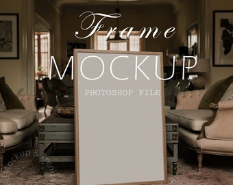 May include: A light brown wooden frame mockup with a blank canvas, sitting on a patterned rug in a living room setting. The frame is leaning against a vintage-style side table with a decorative bowl on top. Two couches are visible in the background, one with a green velvet cushion and the other with a pink cushion. The text "Frame Mockup" and "Photoshop File" is visible on the image.