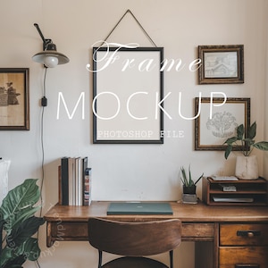 Pode incluir: Uma maquete de moldura preta pendurada em uma parede branca com uma mesa e uma cadeira de madeira na frente. A moldura é cercada por outras obras de arte emolduradas e uma planta. O texto "Frame Mockup" e "Photoshop File" está escrito na parede.