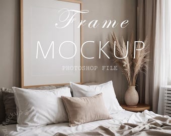 May include: A large, rectangular wooden frame mockup with the text "Frame MOCKUP Photoshop File" is displayed above a bed with white pillows and a beige blanket. A vase of dried pampas grass sits on a small wooden table.