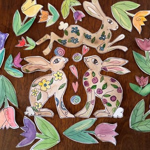 May include: Watercolor illustrations of bunnies and flowers in shades of brown, pink, purple, yellow, and green. The bunnies are decorated with floral patterns. The flowers include tulips and other spring blooms. The artwork is arranged on a wooden surface.