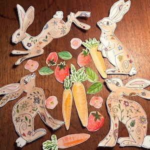 May include: Watercolor illustrations of Easter bunnies, carrots, strawberries, and flowers. The bunnies are brown with floral and swirl patterns. Carrots are orange with green tops, and strawberries are red. The artwork is on a wooden surface.