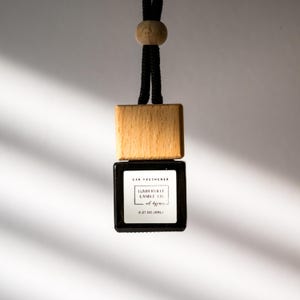 May include: A car air freshener with a wooden top and a black glass container. The label reads "Lumberville Candle Co." and "all seasons". The freshener hangs from a black cord with a wooden bead. The container holds 0.27 oz (8mL) of fragrance.