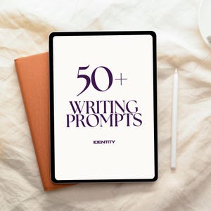 Op de afbeelding: Een digitale tablet toont de tekst "50+ WRITING PROMPTS" in donkerpaars. Een oranje notitieboek en een witte stylus liggen naast de tablet. Het woord "IDENTITY" staat onder de tekst. De achtergrond is een lichtbeige stof.