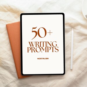 Op de afbeelding: Een digitale tablet toont "50+ WRITING PROMPTS" in bruine tekst, met "NOSTALGIA" eronder. Een witte stylus en een oranje notitieboek liggen naast de tablet. De achtergrond is een crèmekleurige stof.