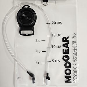 Op de afbeelding: Een doorzichtige waterreservoir met een zwarte dop, slang en bijtventiel. Het reservoir heeft metingen in ponden en liters, met de tekst "MODGEAR WATER WEIGHT 2+" op de voorkant. Bovenop zit een zwarte band.