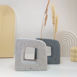 Op de afbeelding: Twee kleine, decoratieve betonnen vazen met een ruw, getextureerd oppervlak. De vaas links is wit en de vaas rechts is grijs. Beide vazen hebben een vierkante opening aan de voorkant.