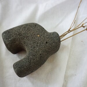 May include: A grey, textured, stone vase with a unique shape. The vase is made of volcanic rock and has a rough, uneven surface.