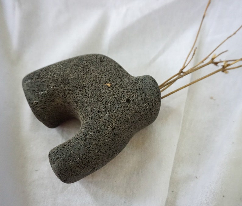 May include: A grey, textured, stone vase with a unique shape. The vase is made of volcanic rock and has a rough, uneven surface.
