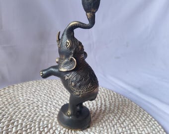 Puede incluir: Un portavelas de elefante de metal negro y dorado. El elefante está de pie sobre sus patas traseras con la trompa levantada, sosteniendo una copa para velas. El portavelas está decorado con intrincados detalles.