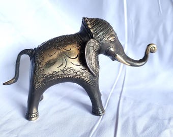 Puede incluir: Una figura de elefante de color bronce con intrincados detalles grabados. El elefante tiene una trompa curvada, orejas grandes y un tocado decorativo. La figura está sobre cuatro patas y tiene una cola curvada.