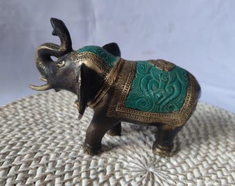 Puede incluir: Una pequeña figura de elefante de bronce con detalles en verde y dorado. La trompa del elefante está levantada y su cabeza está girada hacia un lado. La figura está de pie sobre una superficie blanca tejida.