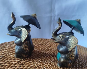 Puede incluir: Dos pequeñas figuritas de elefante de bronce con paraguas. Los elefantes están sentados y tienen las trompas levantadas. Un elefante tiene un paraguas negro y el otro un paraguas verde.