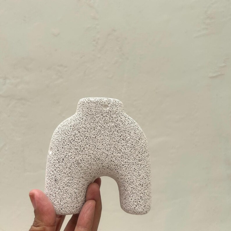 May include: A white ceramic vase with a textured surface and a hole at the top for hanging. The vase is shaped like a human figure with a rounded bottom and a narrow neck.