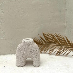 May include: White ceramic vase with a textured surface, shaped like a human figure with legs and a rounded top. The vase is sitting on a white surface with a dried palm leaf behind it.