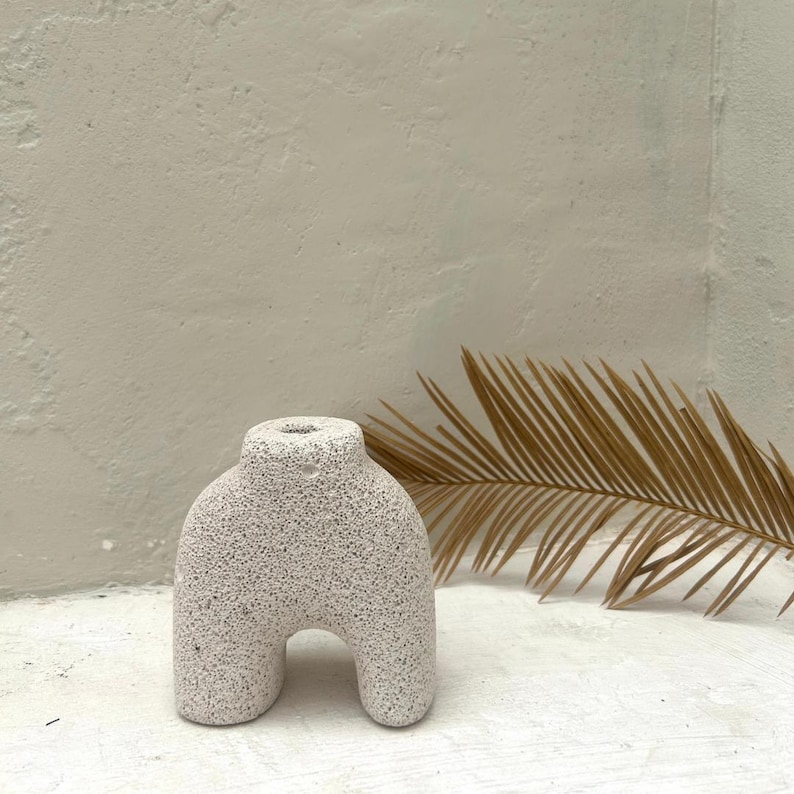 May include: White ceramic vase with a textured surface, shaped like a human figure with legs and a rounded top. The vase is sitting on a white surface with a dried palm leaf behind it.
