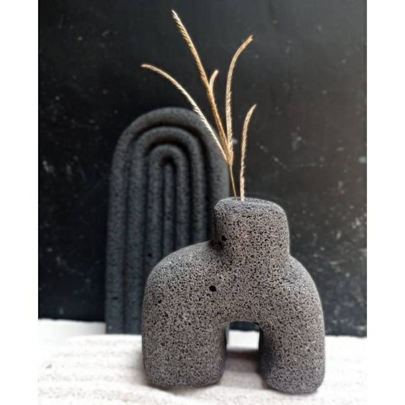 May include: A grey, textured ceramic vase shaped like an arch with a small opening at the top. The vase is sitting on a white surface with dried grasses inside.