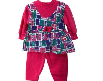 Op de afbeelding: Een roze baby meisjes outfit met een lang mouwen top en broek. De top heeft een wit, groen, roze en paars geometrisch patroon met een strik bij de halslijn.