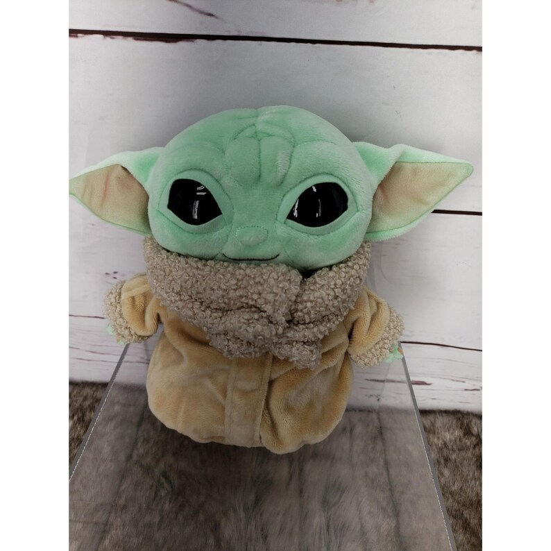 Op de afbeelding: Een pluche speelgoed van een personage met grote zwarte ogen, groene huid en grote oren. Het personage draagt een bruine jas met een fuzzy kraag. Het speelgoed is ongeveer 25 cm hoog.