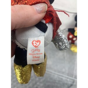 以下が含まれることがあります： 金色のグリッターシューズ、赤いマント、銀色のグリッターアクセントが付いたミニーマウススパークルぬいぐるみ。タグには「Sparkle Minnie Mouse Disney」と書かれています。ぬいぐるみは人が持っています。