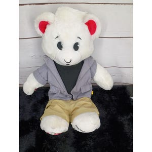May include: A white plush bear with black embroidered eyes and nose, featuring bright red inner ears. The bear wears a gray collared jacket over a black shirt and tan shorts. The word GHOST is printed on the white fabric of the left foot.