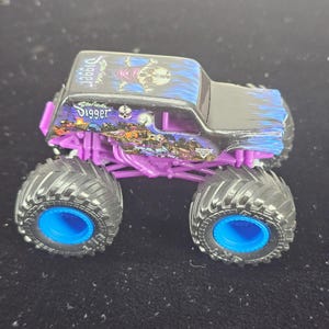 May include: A Son-uva Digger monster truck toy featuring a black body with blue flame graphics, a skull illustration on the roof, and purple chassis components. The vehicle has large black tires with bright blue rims and detailed side decals.