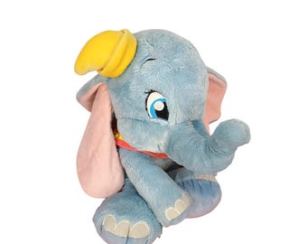 May include: A plush blue elephant toy with large pink ears, a yellow hat, and a red collar. The elephant has large blue eyes and is sitting down. The toy is made of soft material and is designed to look like the character Dumbo.