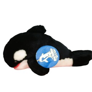 May include: A plush orca whale toy, primarily black with white patches and a red mouth. A blue circular tag is attached to the side. The toy is designed to resemble a killer whale.