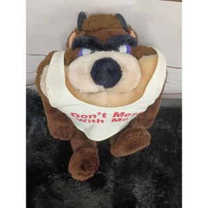 May include: A brown plush toy of the Tasmanian Devil character featuring thick black eyebrows, small horns, and a cream-colored tank top. The tank top has the red text "Don't Mess With Me" printed on the front. The plush has a soft, fuzzy texture.
