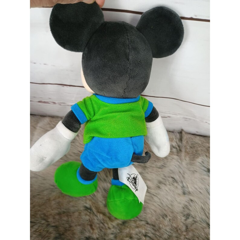 May include: A plush Mickey Mouse doll, viewed from the back, with black ears and head, white gloves, and green and blue clothing. The doll has a tag attached to its foot. The background is a light-colored wooden surface.