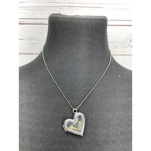 Puede incluir: Un collar de plata con un dije en forma de corazón y una ventana de vidrio transparente que revela una pequeña cruz dorada y otros encantos en el interior.