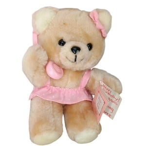 Può includere: Un orsacchiotto di peluche marrone chiaro, che indossa un vestito rosa a pois bianchi e un fiocco rosa. L'orso tiene in mano una cornetta telefonica rosa e un biglietto con le parole "Happy Birthday!" e "Born To Talk".
