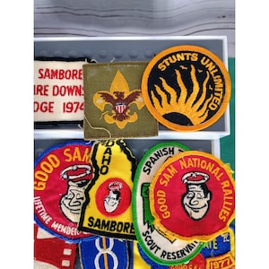 May include: A collection of vintage Boy Scout patches, including a green patch with a scout emblem, a yellow patch with the text "Stunts Unlimited", a red patch with the text "Good Sam", and a yellow patch with the text "Idaho".