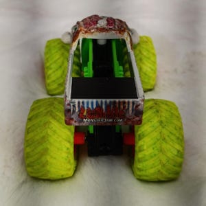 May include: A toy monster truck with a zombie-themed design. The truck features a white and red cab with the text "Grave Digger" and "MonsterJam.com". It has bright green suspension and large, lime green tires.