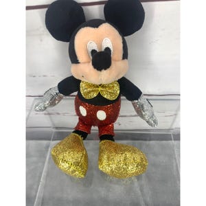 以下が含まれることがあります： 黒い耳、黒い体、ベージュの顔をしたミッキーマウスのぬいぐるみ。おもちゃは白い水玉模様の赤いショートパンツ、銀色の手袋、金色のキラキラ光る靴と蝶ネクタイをしています。おもちゃは透明な表面に立っています。