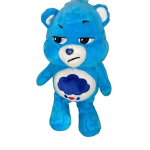 May include: A bright blue plush bear toy with a white muzzle and inner ears. The bear has a grumpy facial expression with embroidered black eyes and a mouth. A white circular patch on the belly features a dark blue rain cloud with red raindrops.