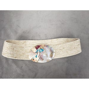 Pode incluir: Um cinto bege tecido com uma fivela oval de concha iridescente. A fivela é adornada com pequenas conchas e detalhes coloridos em azul, rosa e branco. O cinto é de estilo largo, adequado para usar na cintura ou nos quadris.