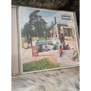 Può includere: Una custodia per CD con la copertina dell'album degli Oasis. L'artwork mostra un orologio, un'auto d'epoca, uno scooter e due uomini. Il nome della band, "Oasis", è visualizzato in un rettangolo bianco in alto.