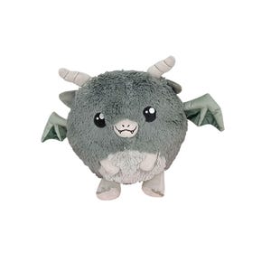 Puede incluir: Un peluche con forma de dragón redondo y gris, con pequeños cuernos y alas. Tiene una barriga y pies de color gris claro, ojos negros y una pequeña boca sonriente. Las alas son de color verde apagado.