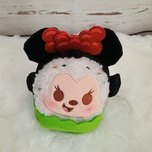 Pu&ograve; includere: Un peluche a forma di personaggio dei cartoni animati, con orecchie nere e un fiocco rosso. Il personaggio ha un viso bianco, guance rosa, occhi marroni e una base verde. Il giocattolo &egrave; ispirato a un personaggio popolare.
