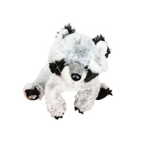 Puede incluir: Un peluche de mapache con pelaje suave moteado en gris y blanco, parches negros alrededor de los ojos y nariz negra. El animal de peluche tiene patas negras, con un pequeño emblema bordado y colorido visible en la planta de una pata.