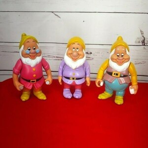 May include: Three plastic dwarf figurines with white beards and yellow hats. The left figure wears a pink tunic, the centre figure wears a light purple tunic, and the right figure wears a tan vest over a blue tunic, all with black belts.