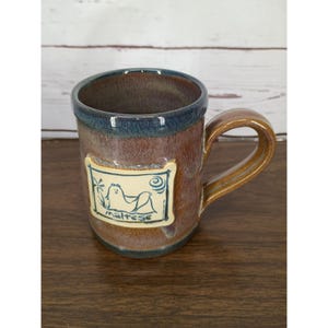 May include: A ceramic mug with a brown and blue glazed finish featuring a rectangular cream-colored plaque on the front. The plaque displays a blue line drawing of a Maltese dog with the text "maltese" written underneath the illustration.
