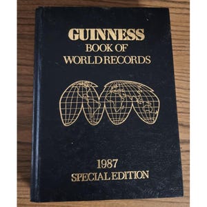 May include: A black hardcover book titled "Guinness Book of World Records" with gold lettering and a gold graphic of interconnected world maps. The book is a 1987 Special Edition.