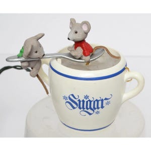 May include: A ceramic teacup with a blue rim and the word "Sugar" in blue script. Two small mouse figurines are perched on a spoon, one wearing a red shirt. The teacup is on a white saucer.