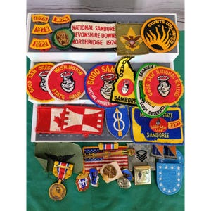 Könnte beinhalten: Eine Sammlung von gestickten Vintage-Pfadfinderaufnähern und Medaillen, angeordnet in einer weißen Schaukasten. Die Stücke weisen verschiedene Formen, Farben und Texte auf, darunter National Samboree, Washington State Rally und Stunts Unlimited.