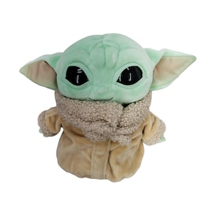Op de afbeelding: Een pluche speelgoed van een personage met grote zwarte ogen, grote oren en een lichtgroene teint. Het personage draagt een bruine jas met een harige kraag. Het speelgoed is een populair personage uit een sciencefictionreeks.