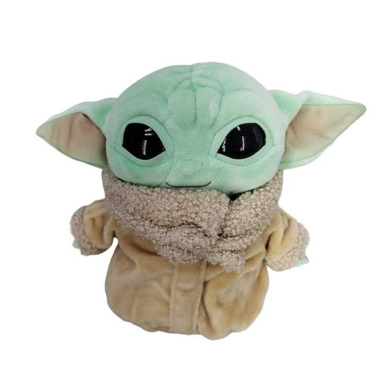 Op de afbeelding: Een pluche speelgoed van een personage met grote zwarte ogen, grote oren en een lichtgroene teint. Het personage draagt een bruine jas met een harige kraag. Het speelgoed is een populair personage uit een sciencefictionreeks.
