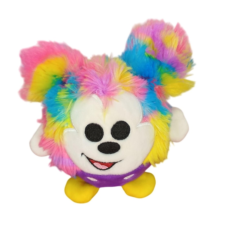 Puede incluir: Un peluche con un personaje de dibujos animados con una cara blanca, ojos negros y una sonrisa roja. El juguete tiene orejas y cuerpo de pelaje esponjoso de color arco&iacute;ris. Tiene manos blancas, una parte inferior morada y pies amarillos.