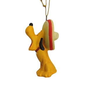 Könnte beinhalten: Gelbe Pluto-Figur mit Sombrero mit roten und weißen Streifen. Die Figur hat eine schwarze Nase und Ohren sowie einen roten Kragen. Sie hängt an einer goldenen Schnur, wahrscheinlich ein Ornament.