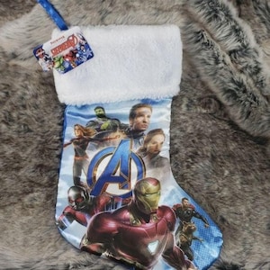 Puede incluir: Una media de Navidad azul y blanca con un puño de piel sintética blanca. La media presenta una imagen impresa de los personajes de los Vengadores, incluidos Iron Man, Capitán América, Thor, Hulk y Viuda Negra. La media tiene el texto "Superhéroe" impreso en el puño.